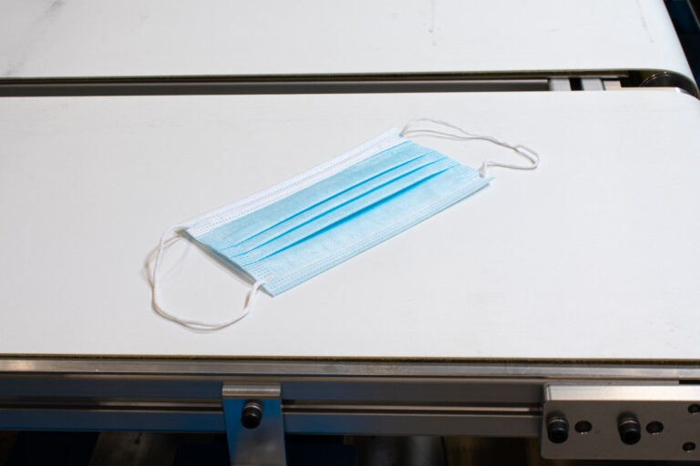 A white table with a blue and white PPE face mask laid diagonally on it with looped strings that attach at the top and bottom and metal trim lining the bottom of the table.