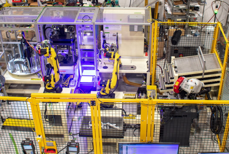 Two yellow robotic arms encased in a black and yellow fenced machine guard with a bright blue light in the middle. Various other components fill the remainder of the fenced in area.