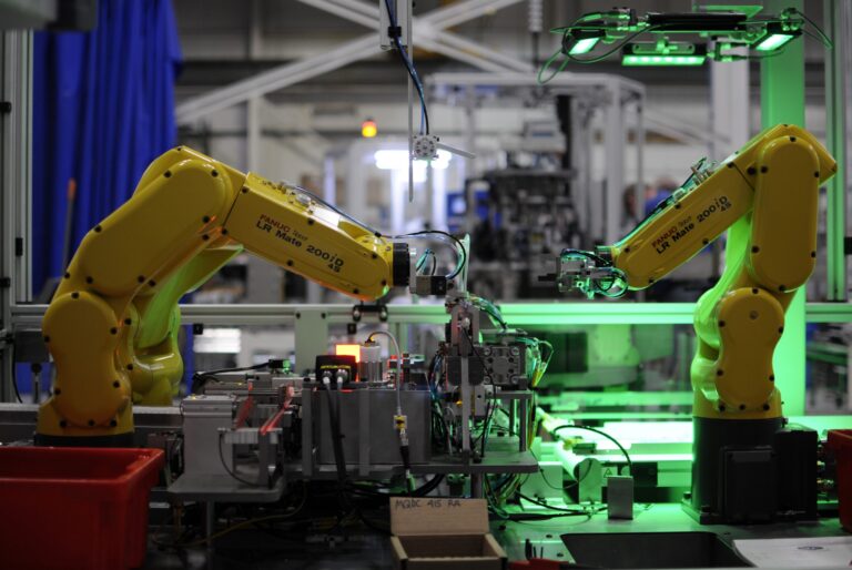Two yellow FANUC robotic arms operating in an industrial automation setting, assembling components on a production line with green lighting and various machinery in the back