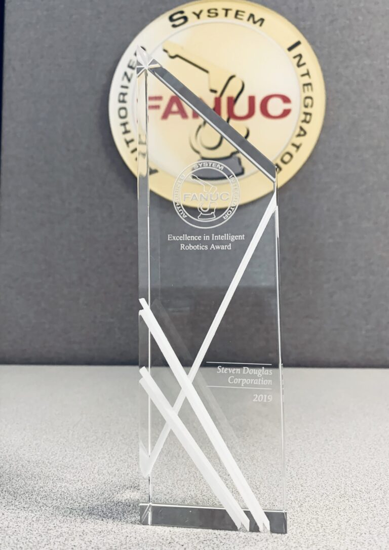 Crystal award with geometric white accents, engraved with 'Excellence in Intelligent Robotics Award' for Steven Douglas Corporation, 2019, against a FANUC logo in the back.