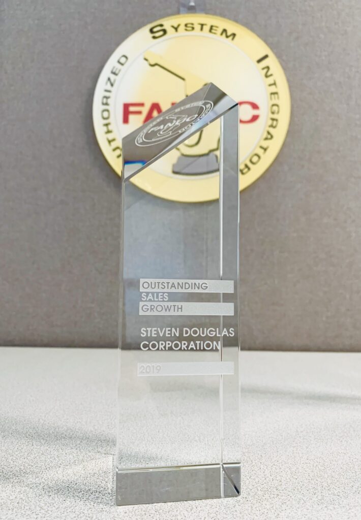 A clear, glass award with an angled top, engraved with "Outstanding Sales Growth - Steven Douglas Corporation - 2019," placed on a desk with a FANUC sign in the background.