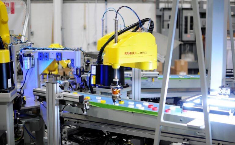 Yellow FANUC SR-6iA industrial robotic arm operating on an automated assembly line, handling colored bottle caps in a high-tech manufacturing facility.