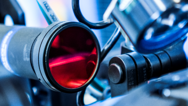 Close-up of an industrial camera with a red lens, surrounded by cables and mechanical components, capturing precision in automation and manufacturing environments.