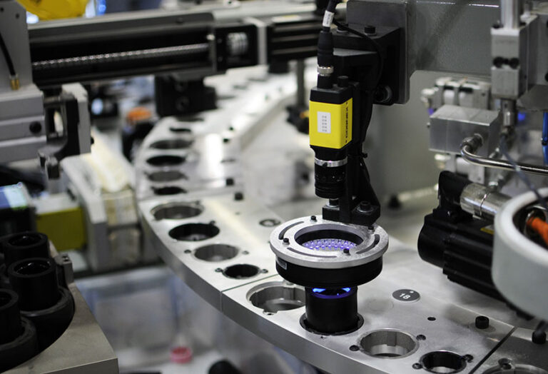 Automated assembly line with robotic inspection system. Vision camera with yellow housing is mounted above a rotating platform, inspecting a component.