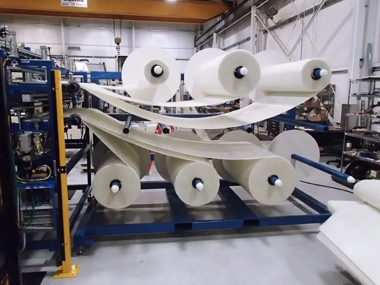 A machine unwinding multiple large rolls of plastic film in an industrial setting, with blue rollers guiding the material through the process.