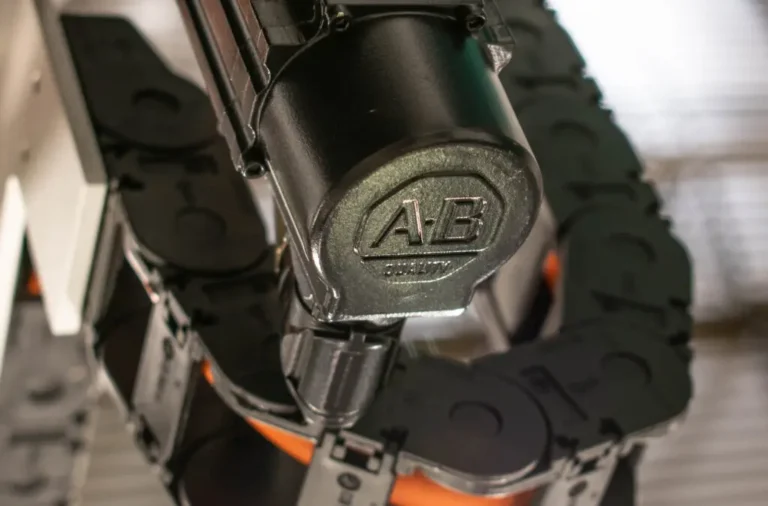 Close-up view of an industrial automation component with "A-B Quality" embossed on the surface, surrounded by black cable chains and mechanical parts.