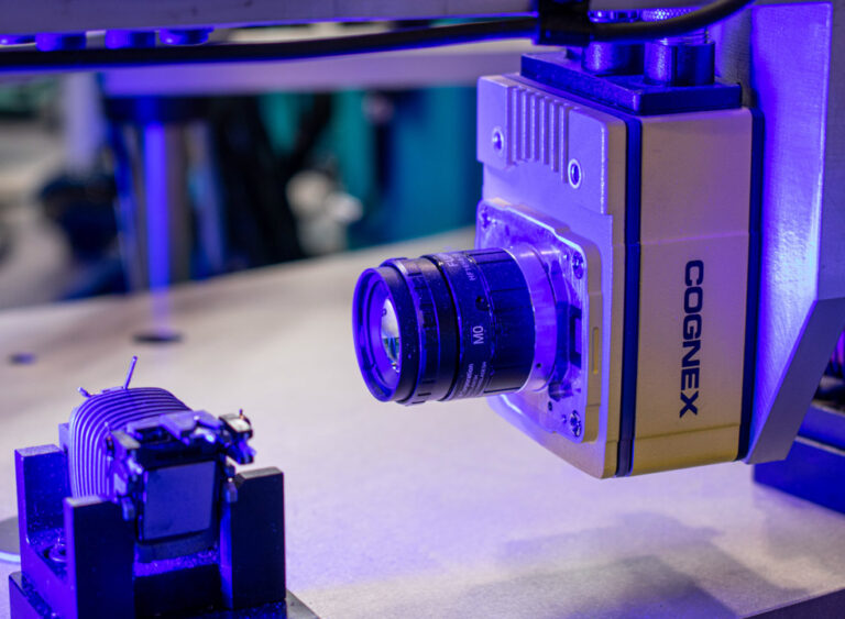 Close up of a yellow Cognex camera inspecting a part that is being used for the automotive industry. Blue lighting is illuminating both the part as well as the camera.