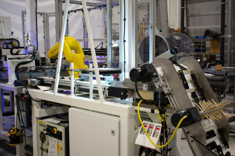 Automated assembly line featuring a yellow robotic arm placing components on a conveyor belt. Various machines line up, including equipment for feeding and processing parts.