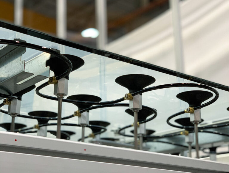 Series of black mounts are attached to a sheet of glass, held in place by thin metal rods and supported by a white, sturdy frame. The mounts are connected via small black tubing to a larger apparatus.