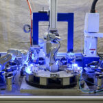 Assembly line features several articulated robotic arms working together. The line incorporates a circular rotating platform with components stationed for processing.