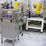 Left of frame is a stainless-steel modular framed storage and a transparent housing. To the right, a yellow robotic arm positioned over a piece of equipment inside a transparent housing.