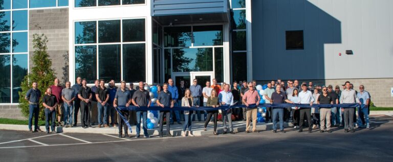 A group of people stands in front of a modern, two-story building with large windows, posing for a traditional ribbon-cutting ceremony. They hold up a wide, dark blue ribbon, poised to cut it.