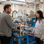 In a well-lit manufacturing facility, two engineers wearing safety glasses are inspecting an automated piece of equipment.