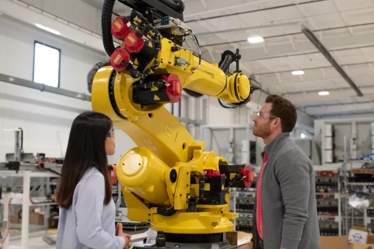 In a brightly lit factory, a man and a woman stand on either side of a large, yellow industrial robot, looking upwards at its articulated arm.