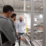 Three people inspect a production line inside a factory with a person holding written drawings in the middle.