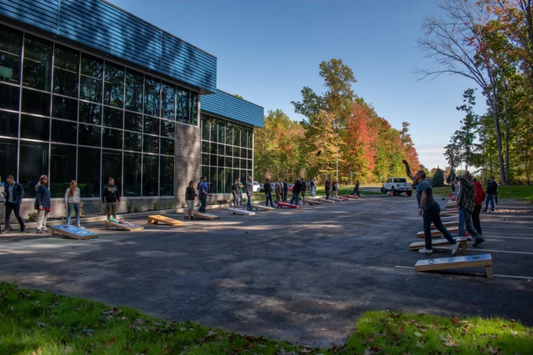 A group of adults plays cornhole in a parking lot next to a large, modern building with mirrored windows. Numerous cornhole boards are arranged in a line across the parking lot.