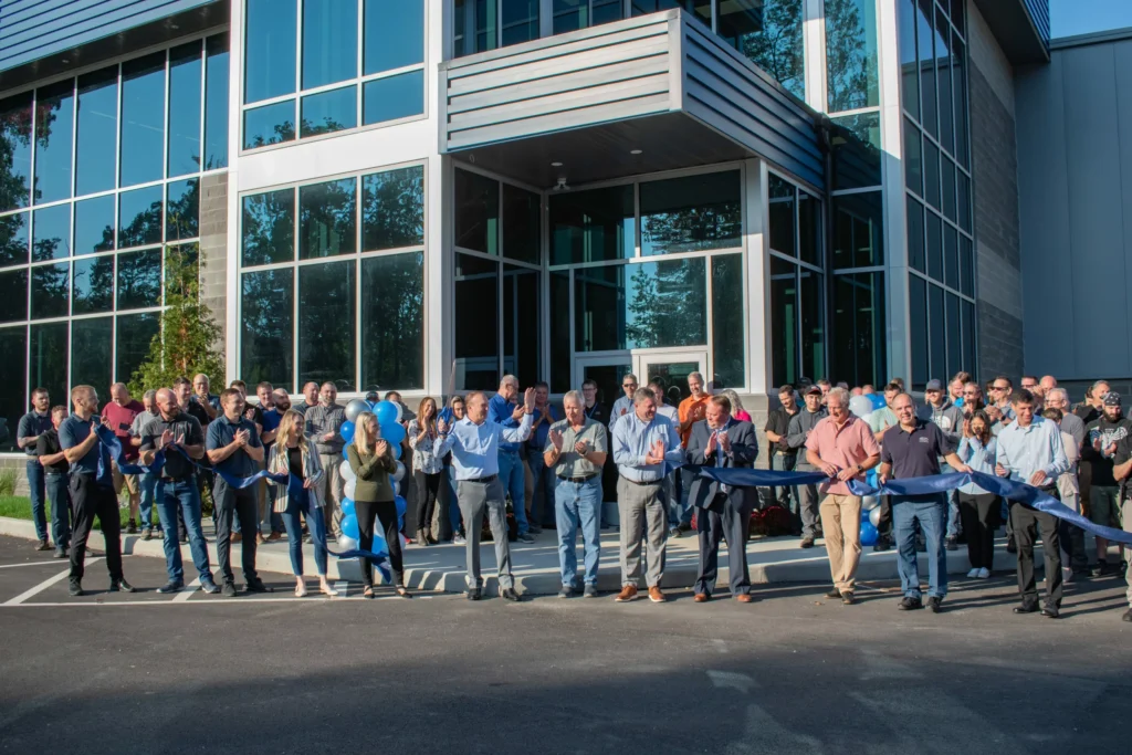 A ribbon-cutting ceremony is taking place in front of an office building. A crowd of people is gathered, with several individuals holding a long blue ribbon and preparing to cut it.