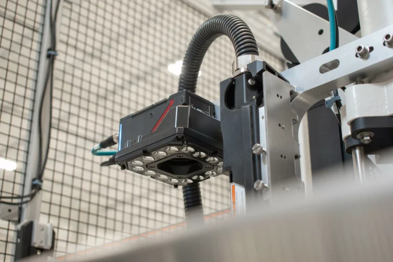 Close-up of an automated industrial imaging system with attached lights, camera, and support structure. The main system is black and silver, connected to a flexible hose with electrical cables.