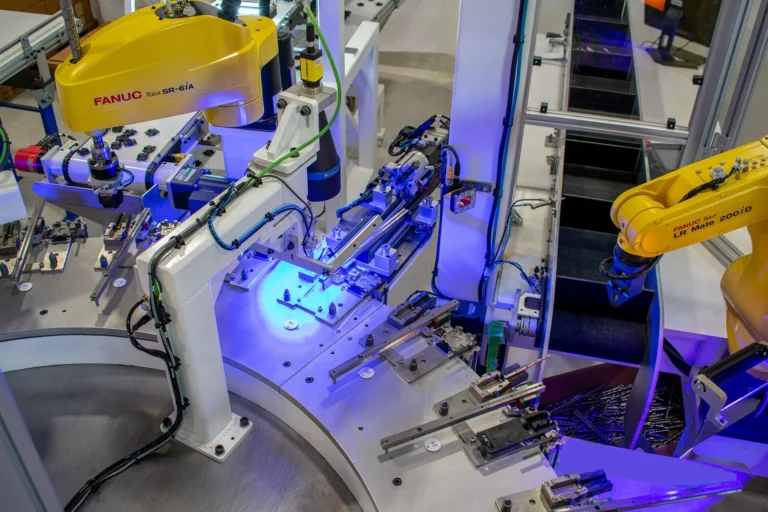 Yellow robotic arm automation system for manufacturing, with blue lights illuminating the workspace and two FANUC robotic arms around rotary table.