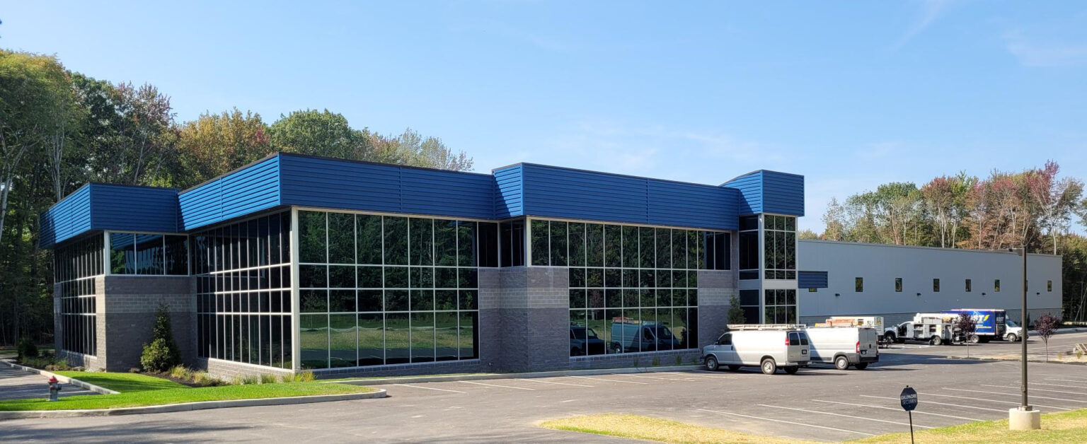 Exterior view of a modern commercial building with a blue roof and gray facade, featuring a large expanse of multi-paned windows. Several white vans are parked in the adjacent parking lot.