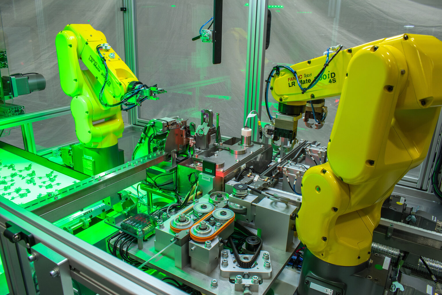 Automated assembly line featuring two bright yellow robotic arms. Green laser beams are visible in the background, part of a sensor or alignment system.