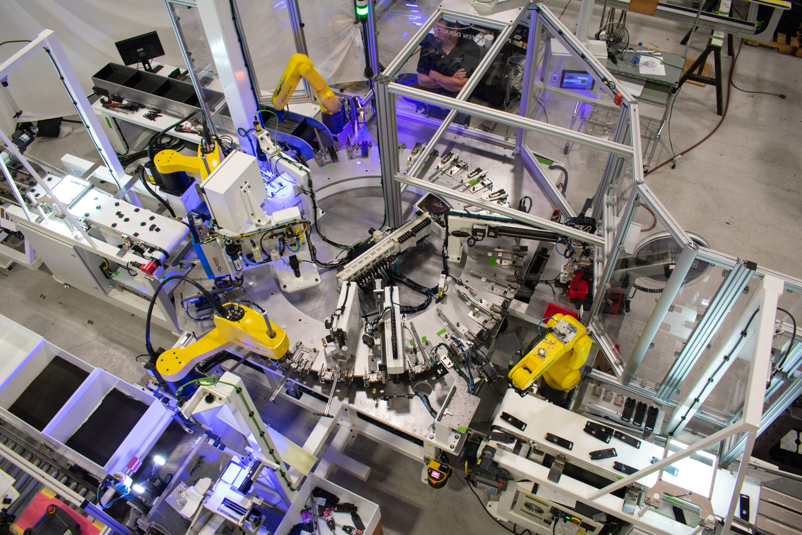 Aerial view of several yellow robotic arms is integrated into the system, manipulating parts on conveyors and within a rotating carousel. Enclosed within clear machine guarding.
