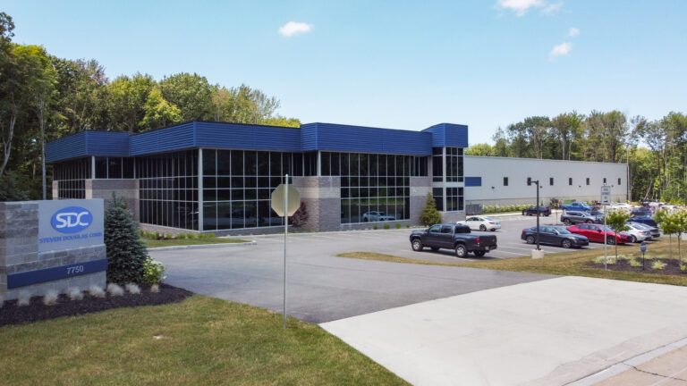 Exterior shot of the SDC Corporation building, a modern industrial structure with blue and grey accents. A parking lot in front of the building is filled with cars.