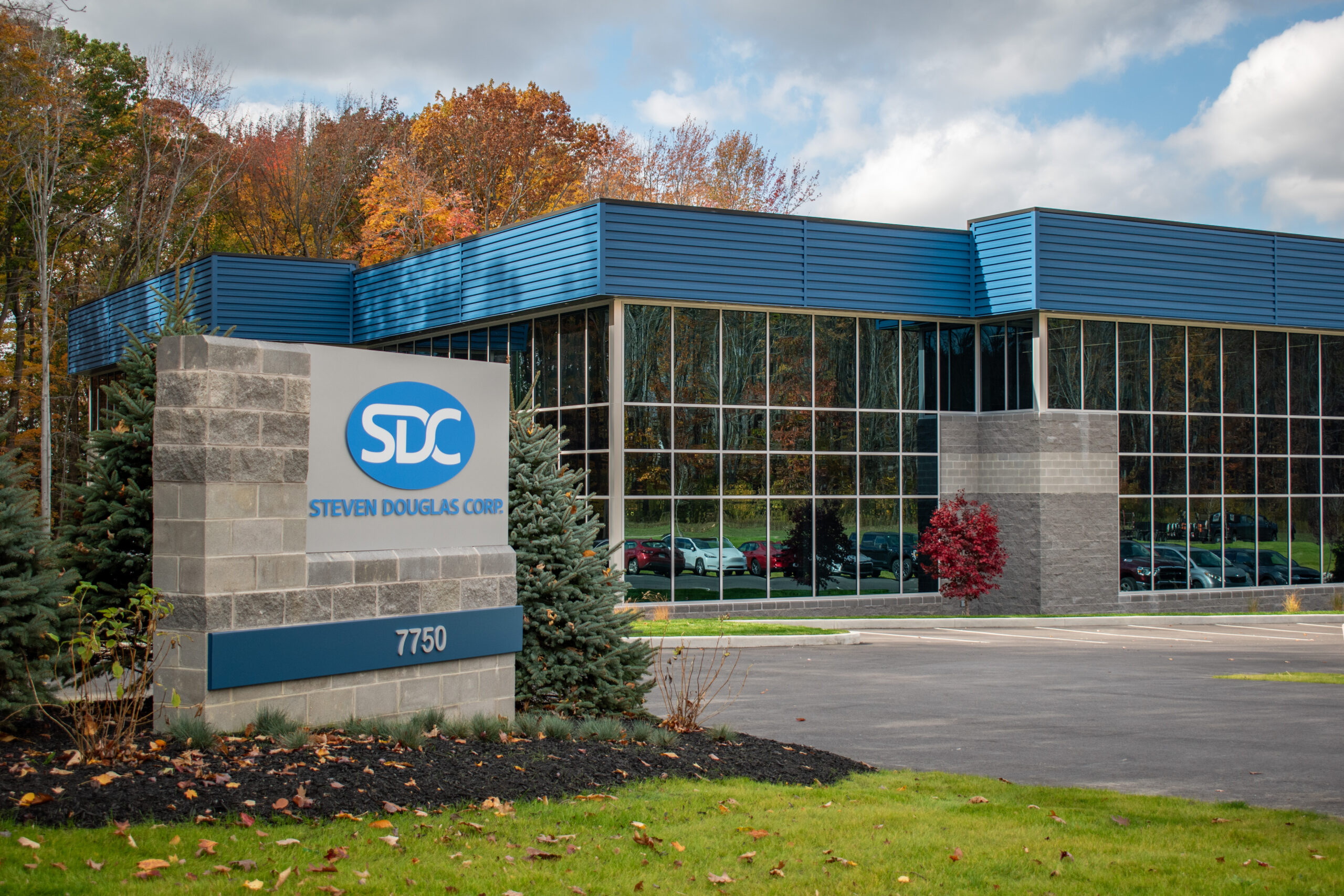 Exterior shot of the Steven Douglas Corporation building. The modern office building features a blue roof and large glass windows. Cars are visible in the parking area.