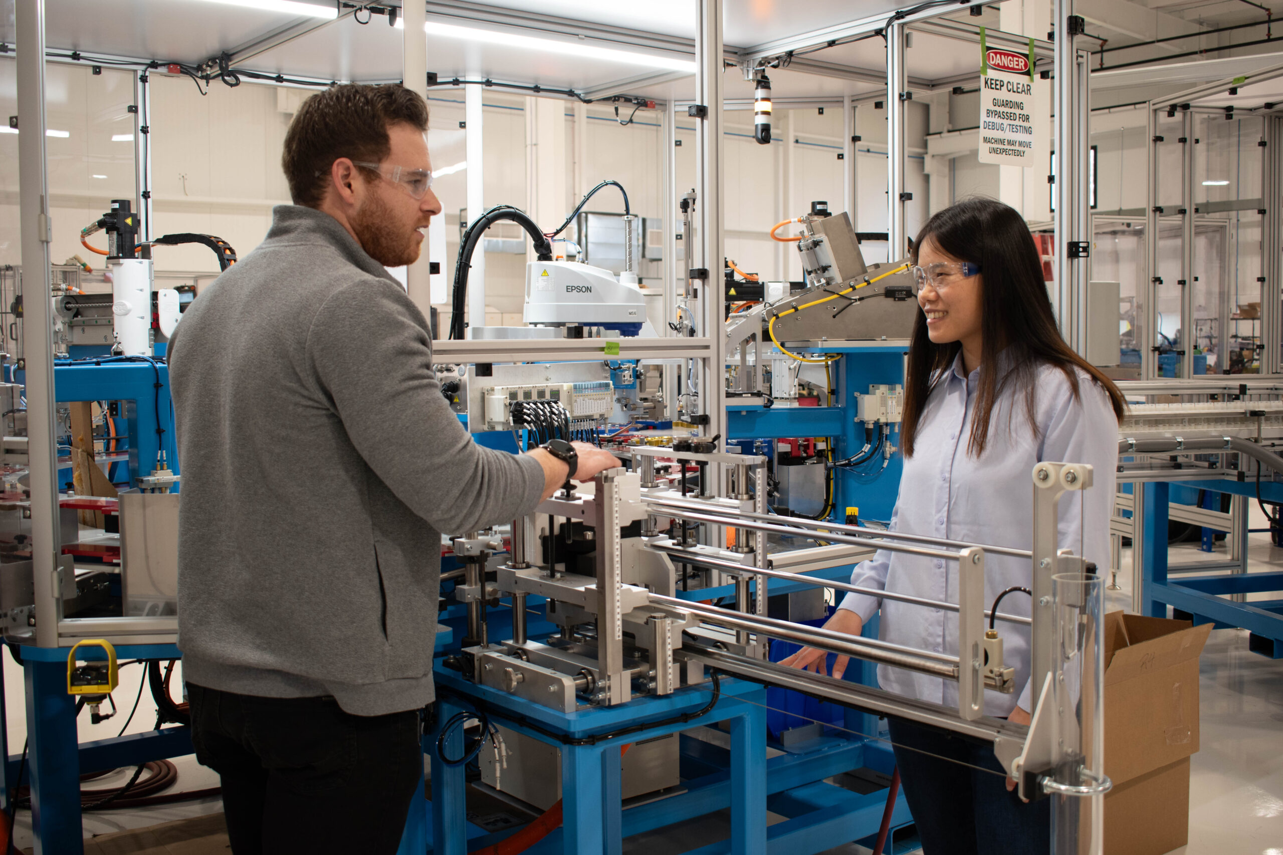 In a factory setting, two engineers are wearing safety glasses are having a conversation while standing near complex machinery and manufacturing equipment.