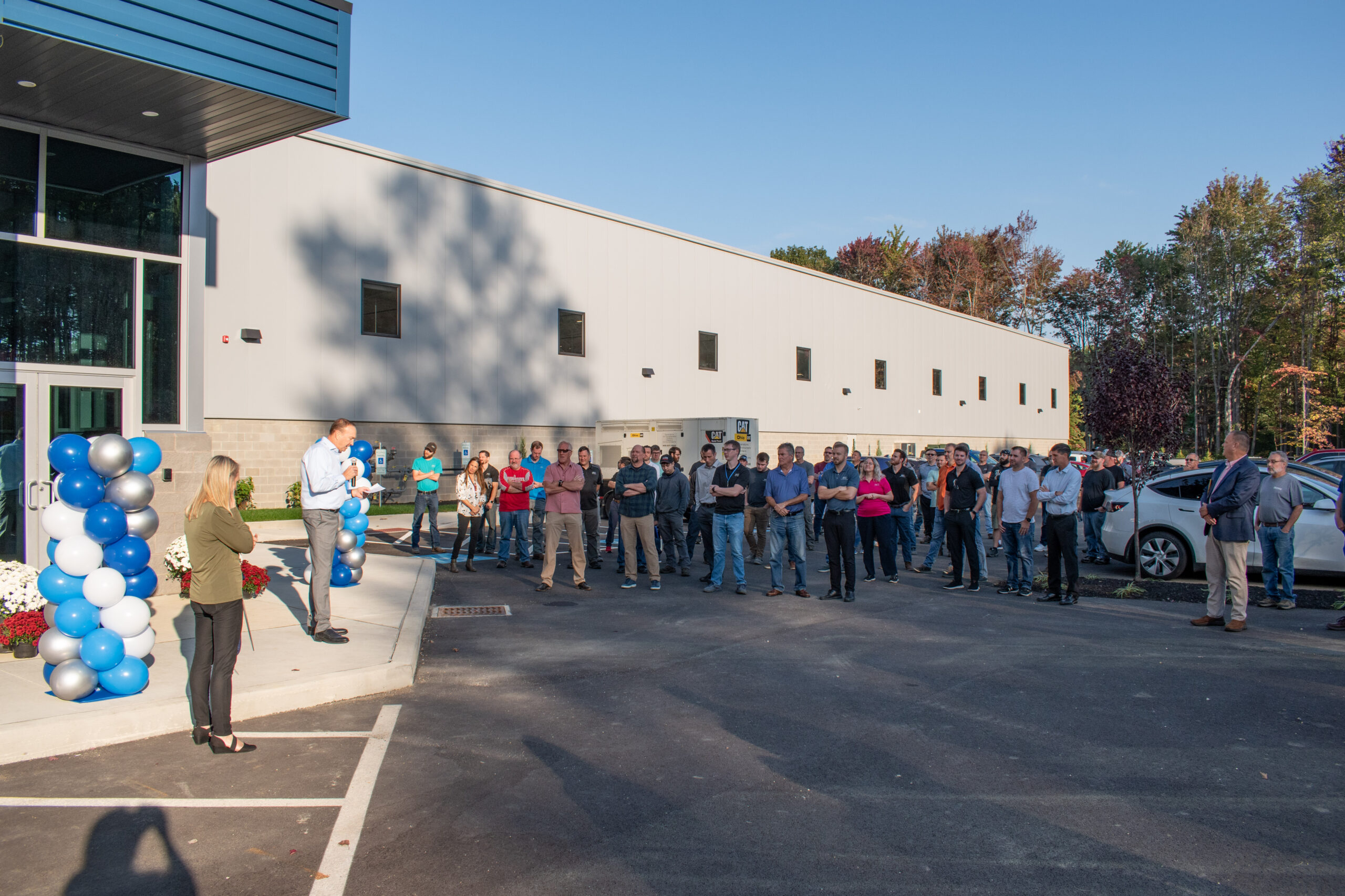 A large group of people stand in front of a newly constructed, modern building, attending an opening or dedication ceremony. A man stands at a makeshift podium, speaking to the crowd.