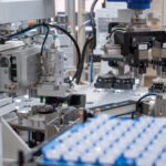 A close-up of an automated assembly machine in a factory setting. The machine features robotic arms and grippers, processing small, clear plastic containers arranged in a tray.