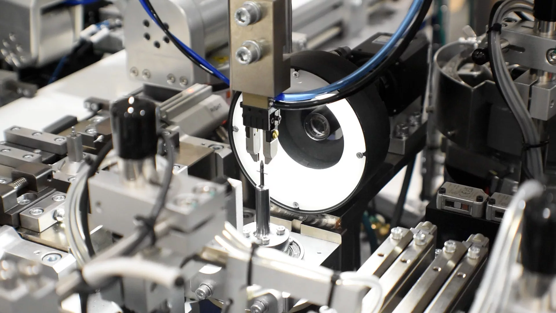Machine is made up of silver metal components of clamps, arms, and sensors. A bright, round backlight illuminates the work area where a process is being performed.