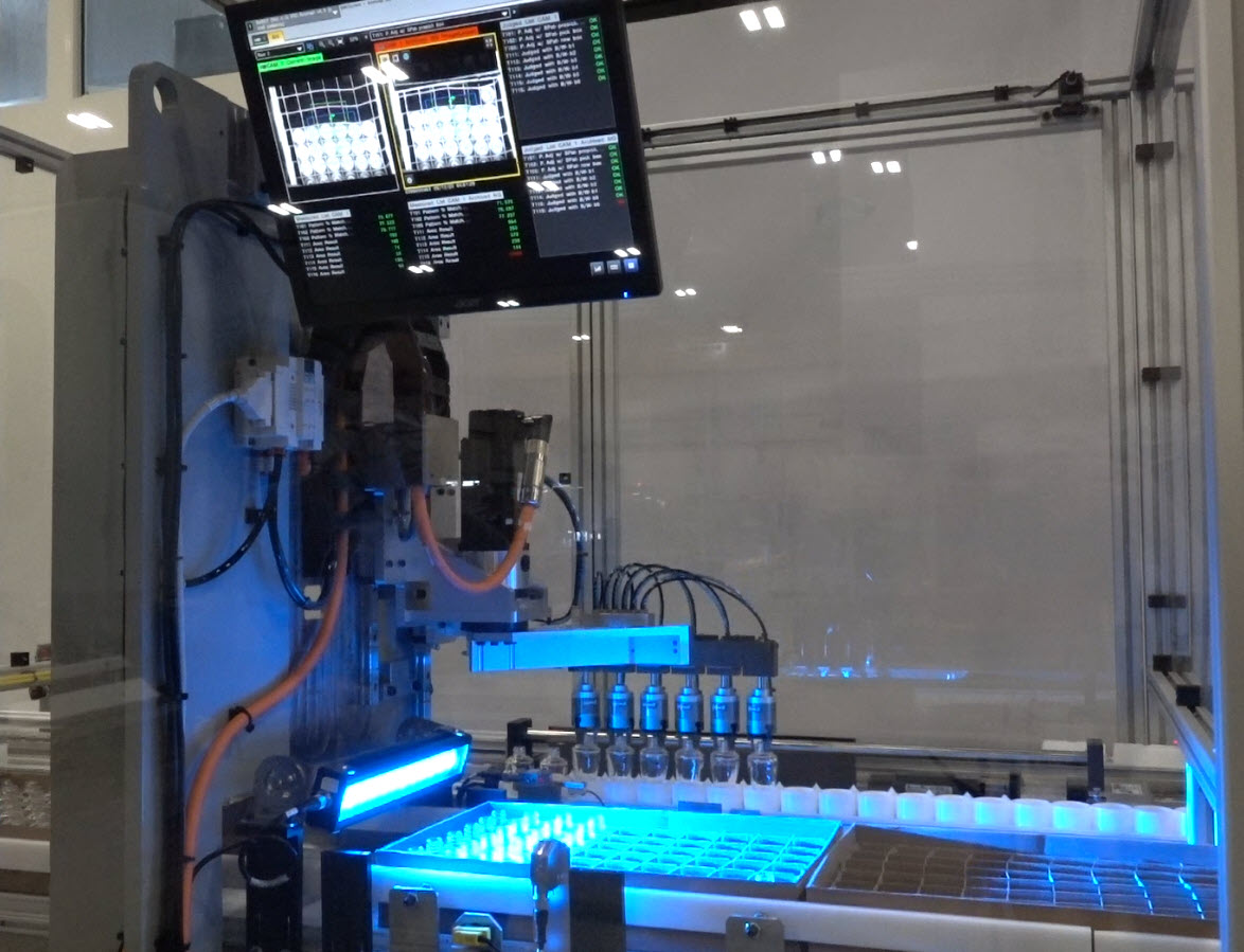 Automated laboratory machine inside a transparent enclosure. The machinery consists of a robotic arm with multiple nozzles positioned over a tray filled with small vials.