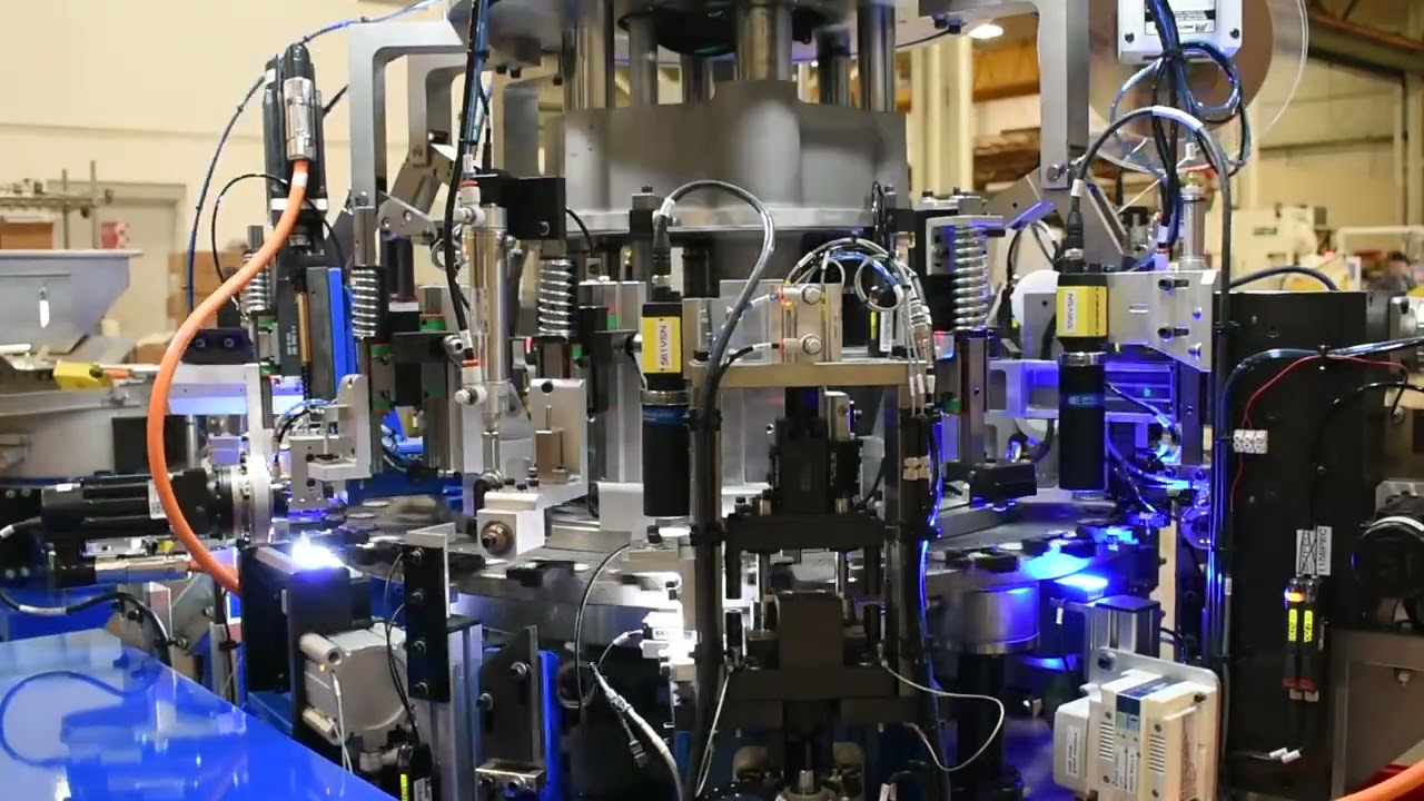 Close-up of a complex automated machine in an industrial setting. The equipment consists of metal framework and numerous sensors, cylinders, and cables.