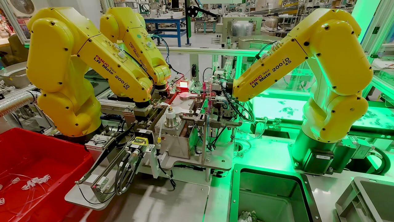 Three yellow FANUC robotic arms are positioned around a conveyor belt platform, assembling small components. Cables extend to a large red bin holding components.