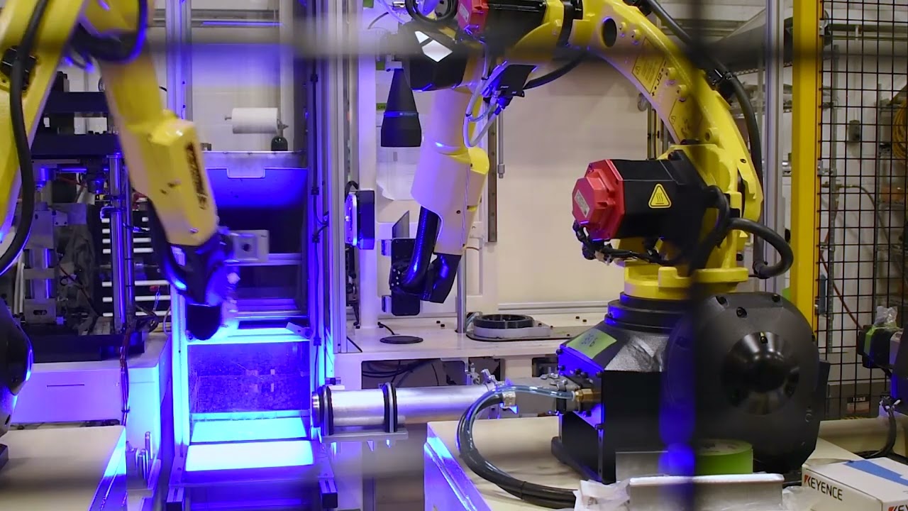 Two yellow robotic arms performing tasks within an automated system. The arms are positioned around a vertical structure, and their movements are highlighted by a strong blue light