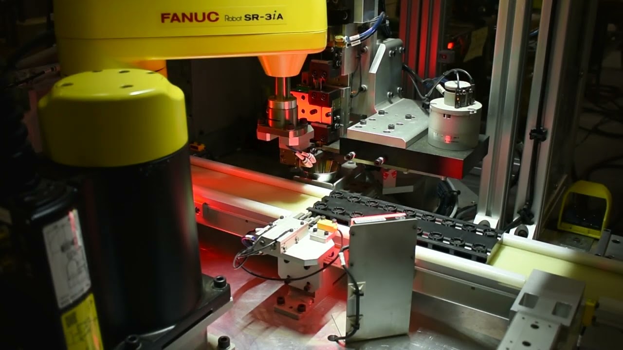 Automated manufacturing assembly line with a yellow FANUC SR-3iA robot arm. The robot is positioned above a conveyor belt, working with precision equipment.