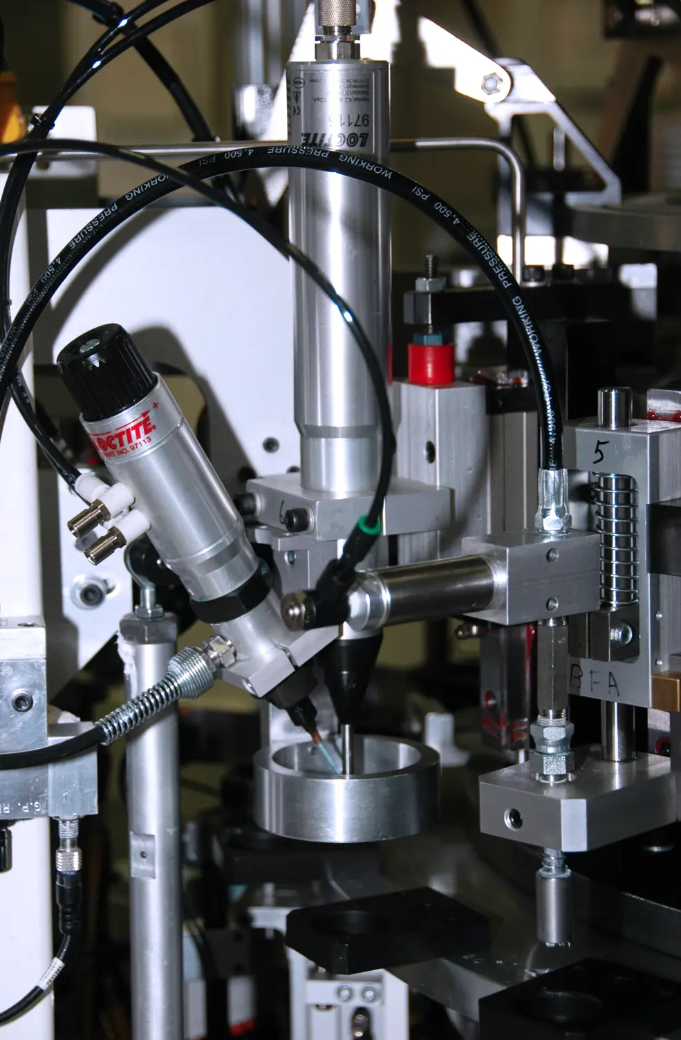 Vertical close-up featuring an automated industrial machine with multiple metal parts. A Loctite dispenser, identified by its label, is angled in the mid-left.