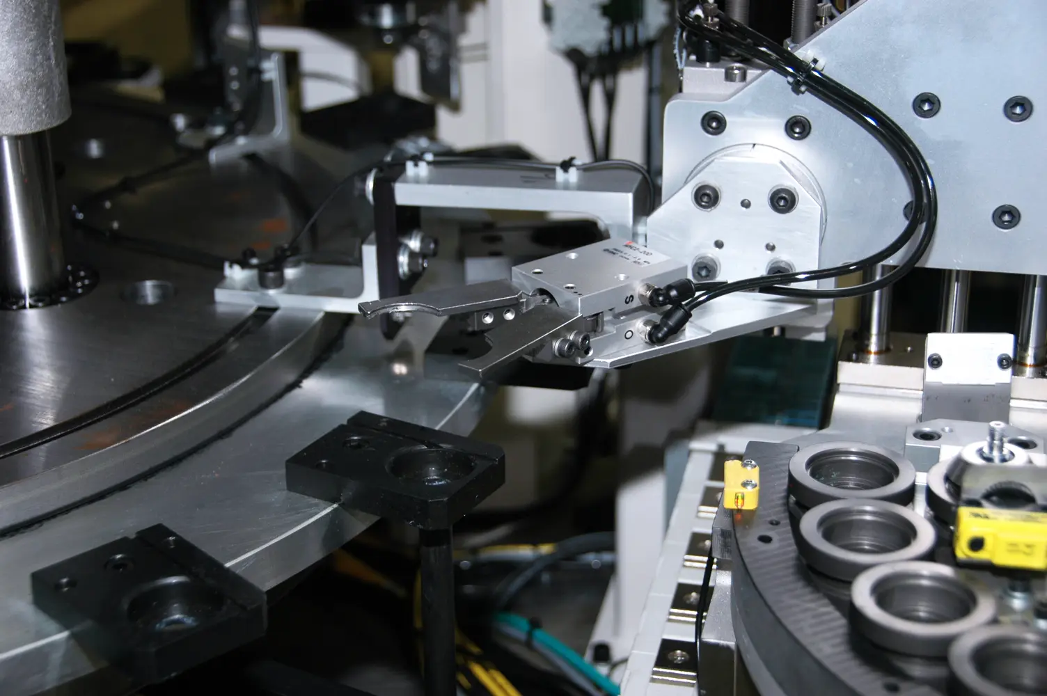 Close-up of an automated industrial machine with a robotic arm. The arm has a pair of tongs/grippers, located above the machine's circular silver colored work surface.