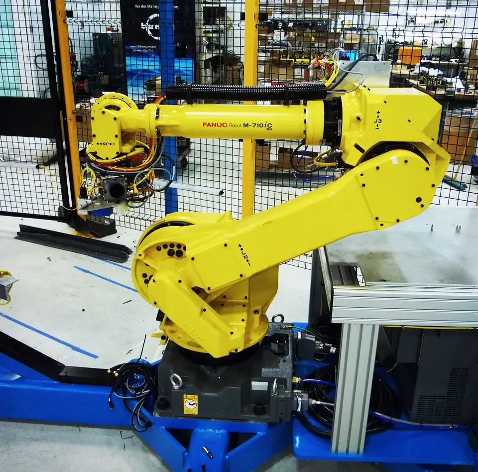 FANUC Robot M-710iC mounted on a black base and inside a fenced machine guard. Connected to wires at the arm and in a bright manufacturing facility.