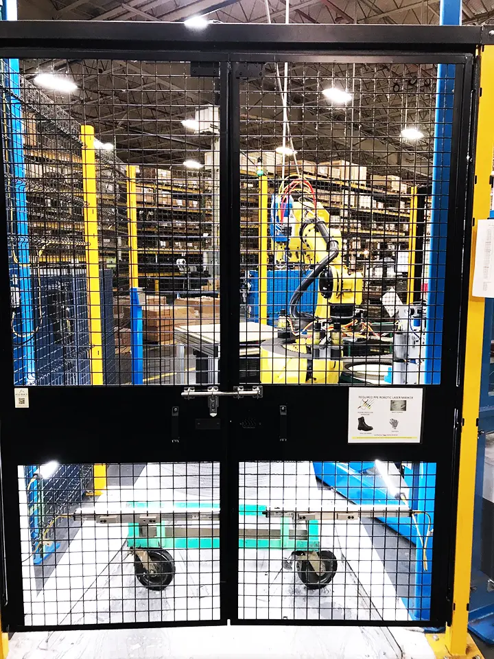 View into a gated-off, yellow-and-blue-framed robotic work cell inside a warehouse. A yellow articulated robot arm is visible inside the enclosure.