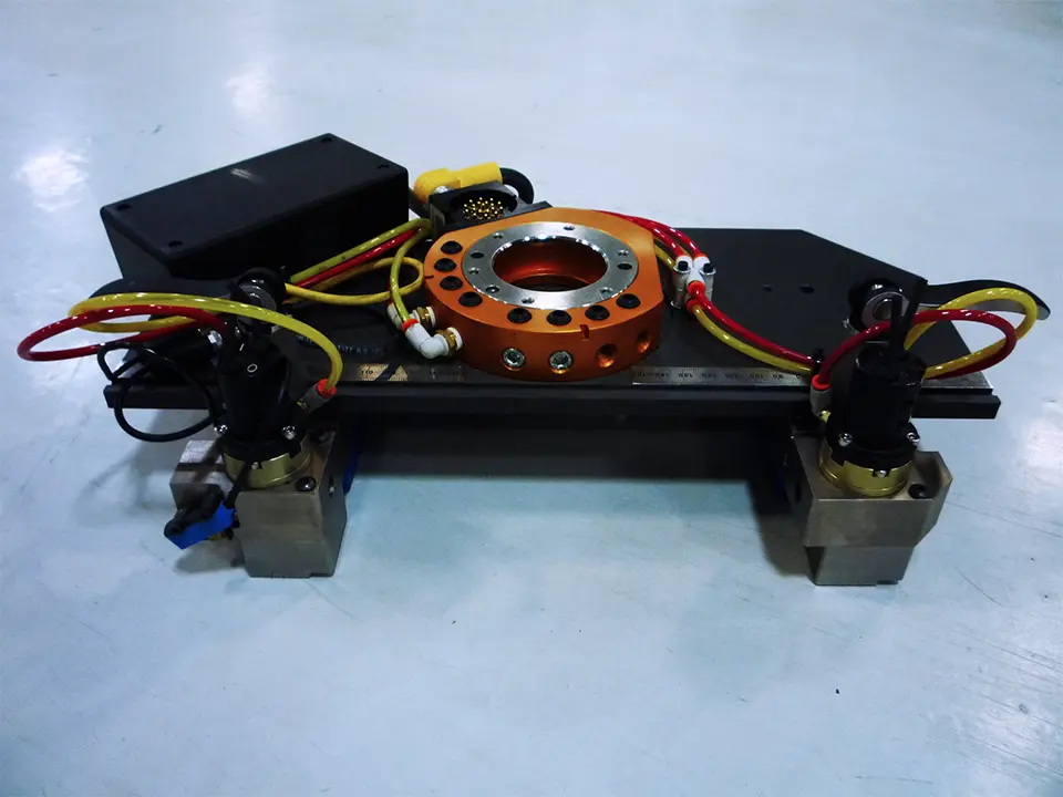 The prototype features an orange circular component with several holes, connected to various hoses and black electronic components.
