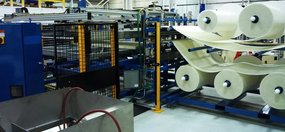 The machinery consists of blue frames and metallic components, automated with a secured safety cage, and a drainage system underneath. The system features large rolls of white material being processed and stacked in multiple layers.