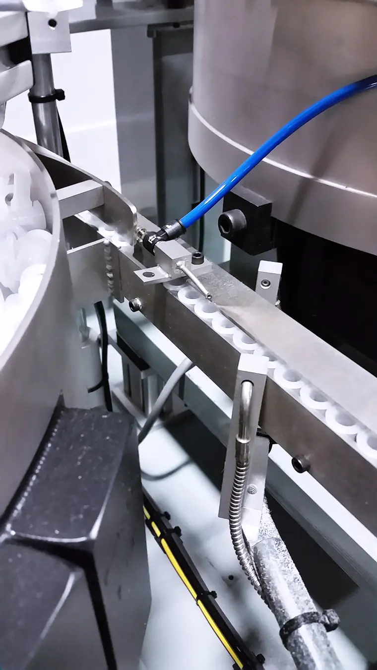 A silver steel rail holds small white plastic rings in evenly spaced recesses, moving them along the line. Blue tubing and other pipes/wires run adjacent to the equipment.