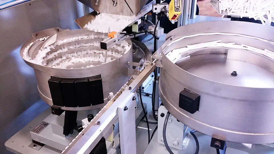 Image shows two automated parts feeders. The left feeder has white pieces circulating in a spiral bowl. The right feeder contains elongated white pieces in its circular tray.