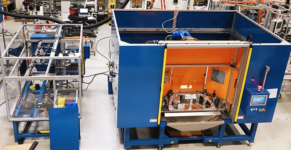 Smaller machine has a silver metal frame and a blue base, and it is for staging. The second, larger blue-and-orange enclosed machine with a user interface on its side appears to conduct the actual assembly process.