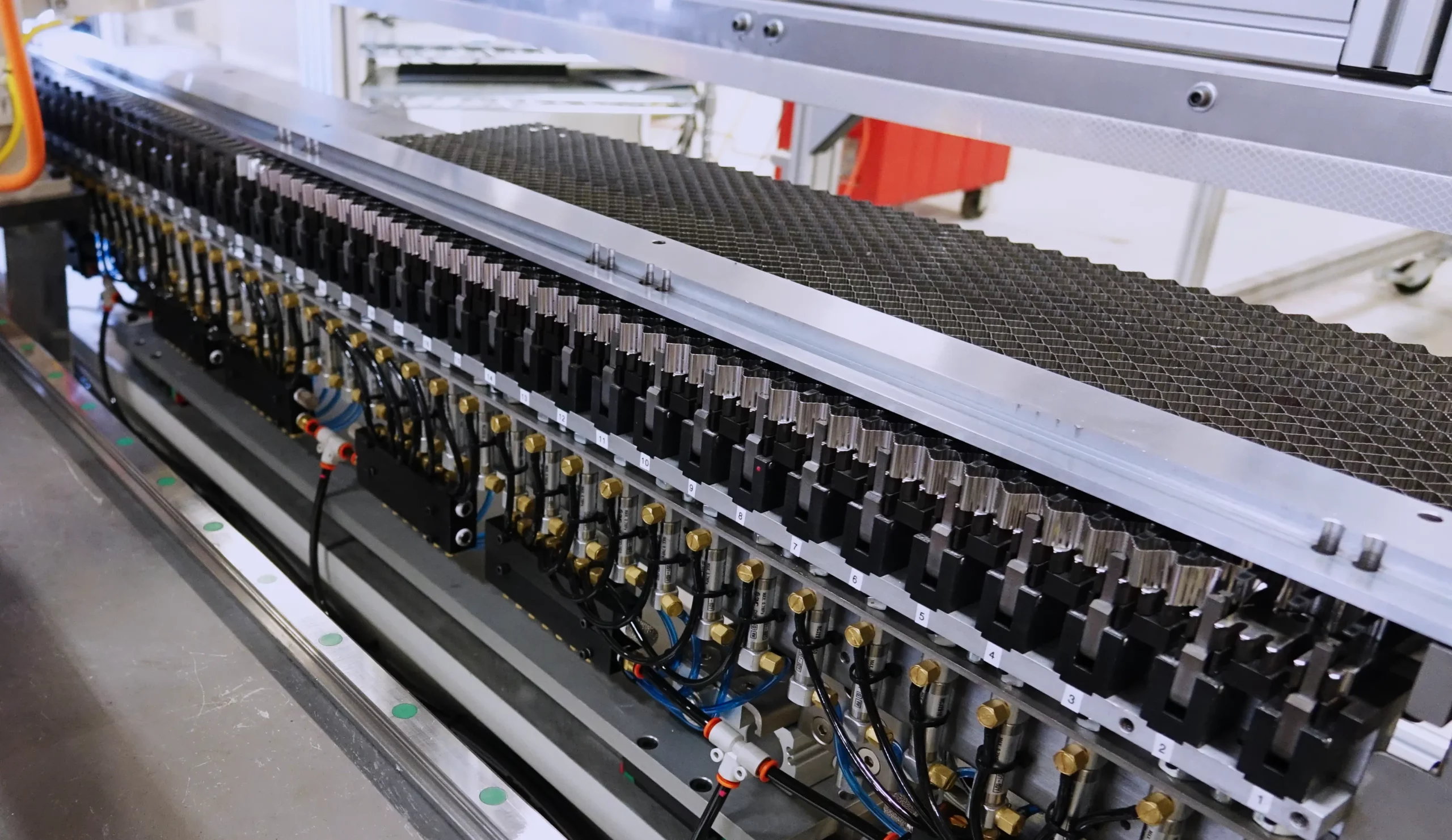 Close-up of a automated system featuring a long row of closely spaced robotic arms positioned above a black honeycomb panel. The machinery is composed of metal and black plastic components, with intricate wiring and hoses connecting the arms.