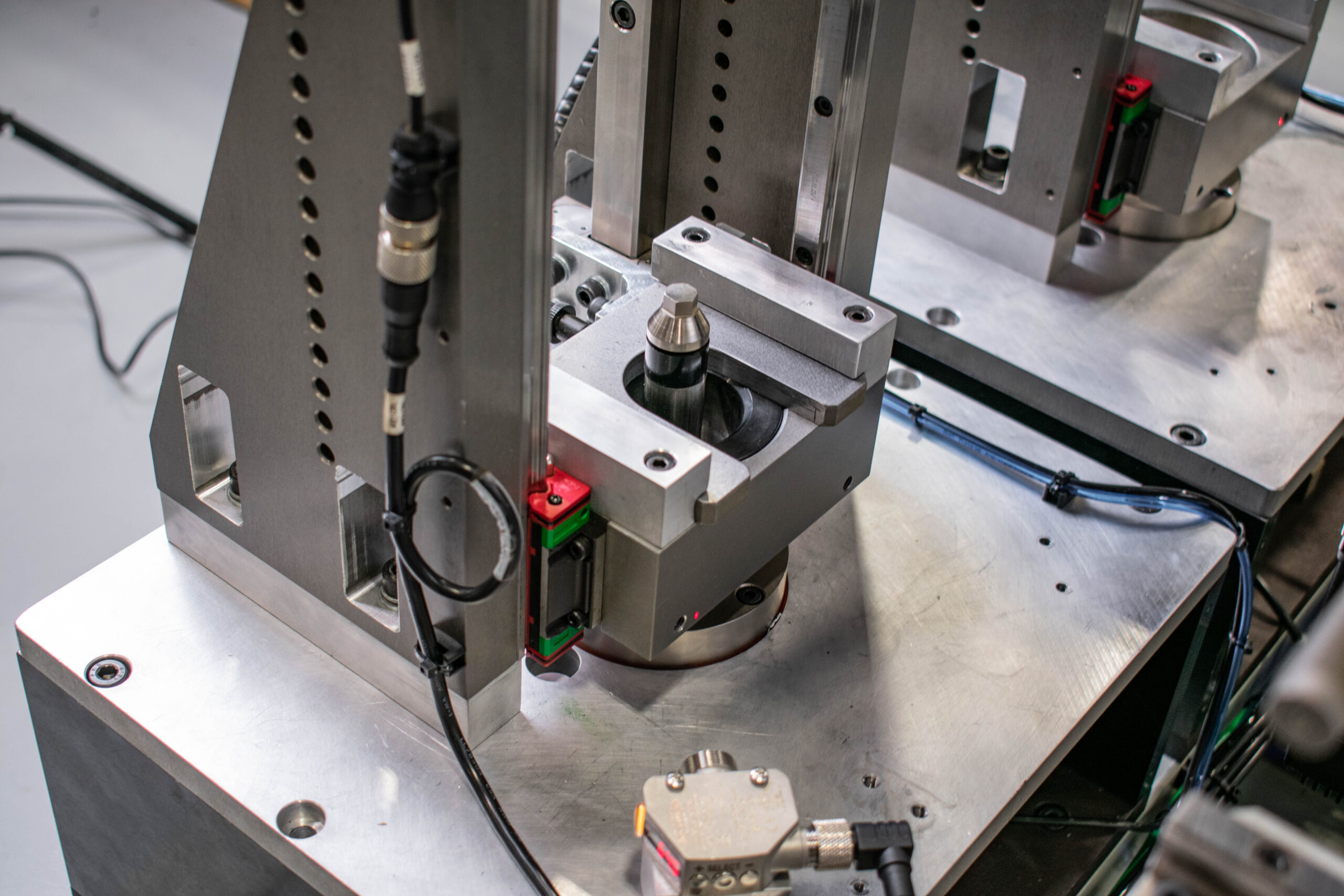 Close-up of an automated industrial machine part during setup. It features a metal component secured in a holding device, with various sensors and cabling visible on a metal base.