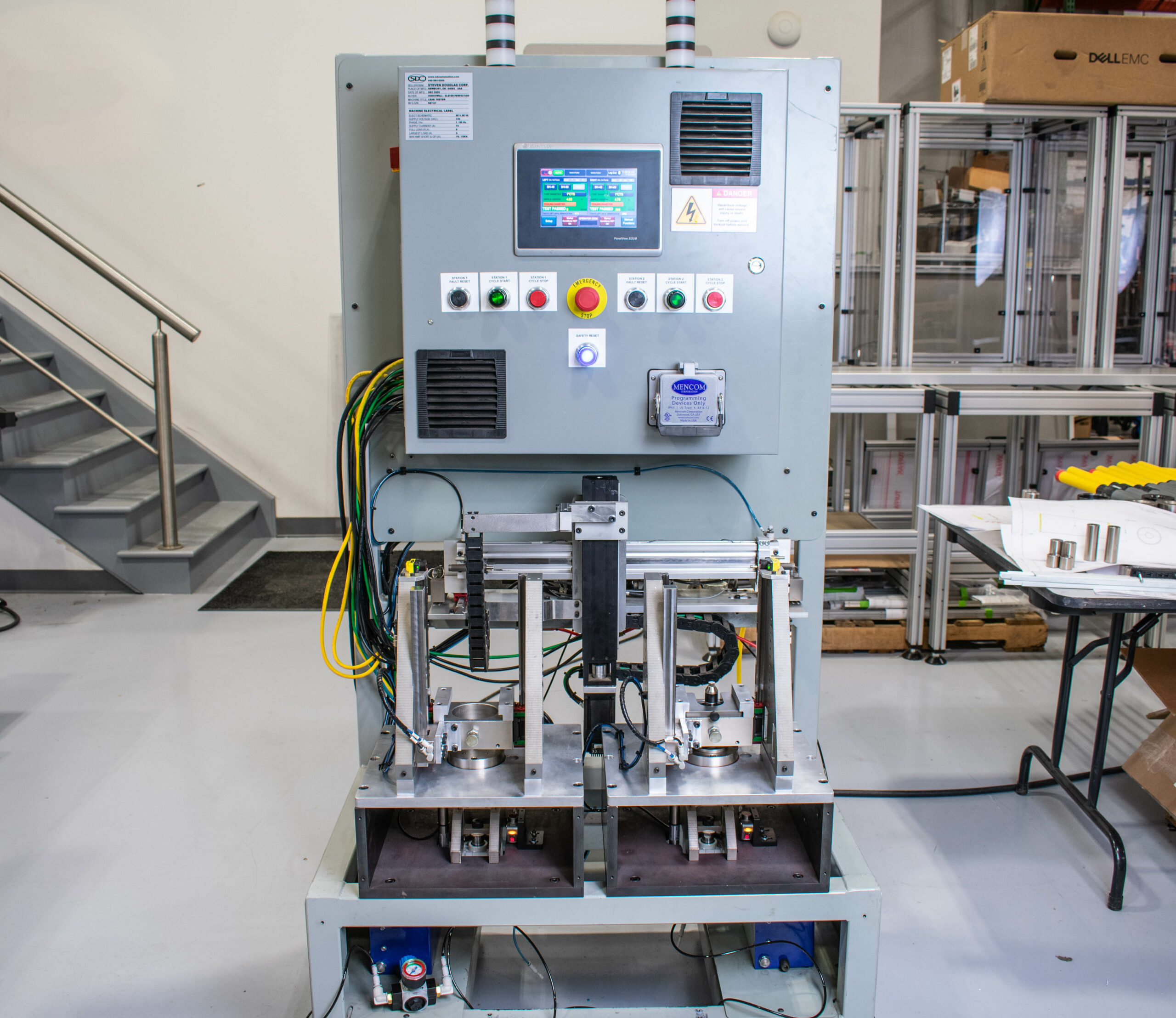A large industrial machine with a gray control panel covered with buttons, indicator lights, a touch-screen control panel displaying graphical data, and a warning sign. Below the panel are two robotic arms poised over metal working stations.