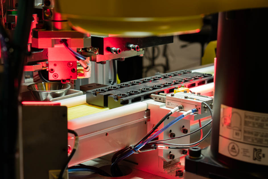 Bearings are arranged in a black tray along a white conveyor belt. A robotic arm with a silver bowl is positioned above the belt, ready to deposit or retrieve components.